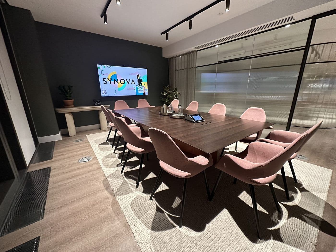Meeting room in a completed Fitzrovia workplace, with a boardroom table, wall-mounted display, Crestron touch panel, track lighting and linear ceiling grilles