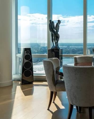 A floorstanding loudspeaker beside floor-to-ceiling glass in a high-rise dining room, positioned discreetly against the window