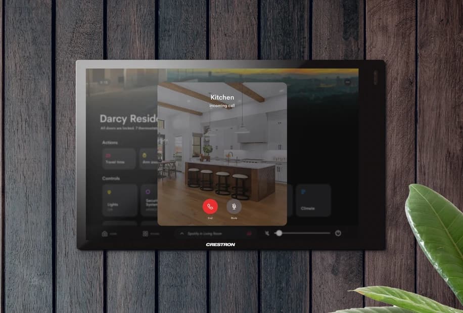 A Crestron touch panel mounted on a dark timber wall showing an incoming call from the kitchen door station, with answer and mute controls
