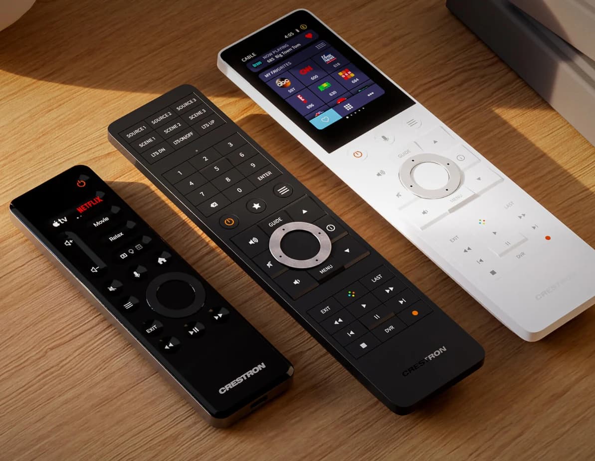 Three Crestron handheld remotes lying on a wooden surface, two black and one white, one with a colour touch screen showing television favourites, the others with engraved scene and source buttons
