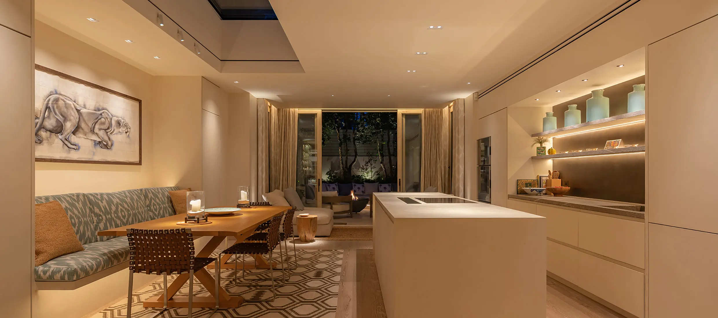 A London kitchen and dining room in the evening: a lit artwork above a banquette, candles on the dining table, concealed cove lighting, lit open shelving, downlights over the island, and the planted garden lit beyond the glass doors